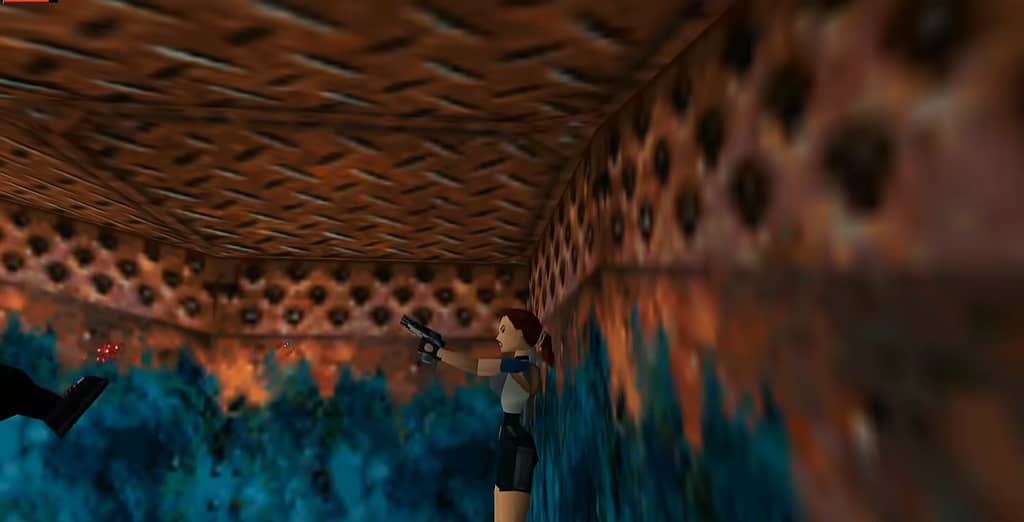 Image descriptor: Within the rusty metal corridors of a sunken cargo ship, Lara Croft points and fires her guns at an enemy off-screen, whose leg can be seen on the left hand side.