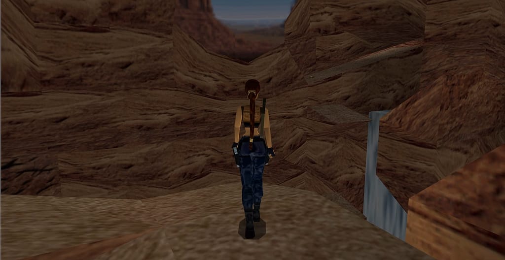 Image descriptor: Lara Croft walks calmly through a giant rocky canyon in Nevada. A waterfall can be seen in the distance on the right hand side, but it is partially obscured by a cliff face's rocks.
