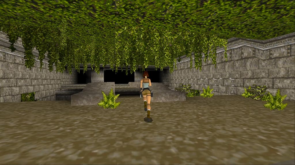 Image descriptor: Lara Croft runs through a Peruvian temple. The floor is made from dirt whilst the walls are made from large stone blocks. The ceiling is grassy and covered in vines, which hang down in the room ahead.